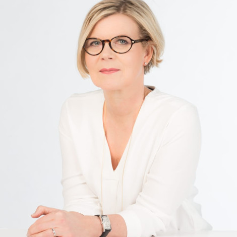 Eeva Kukkosen muotokuva. Hän on pukeutuneena valkoiseen harmaan taustan edessä.